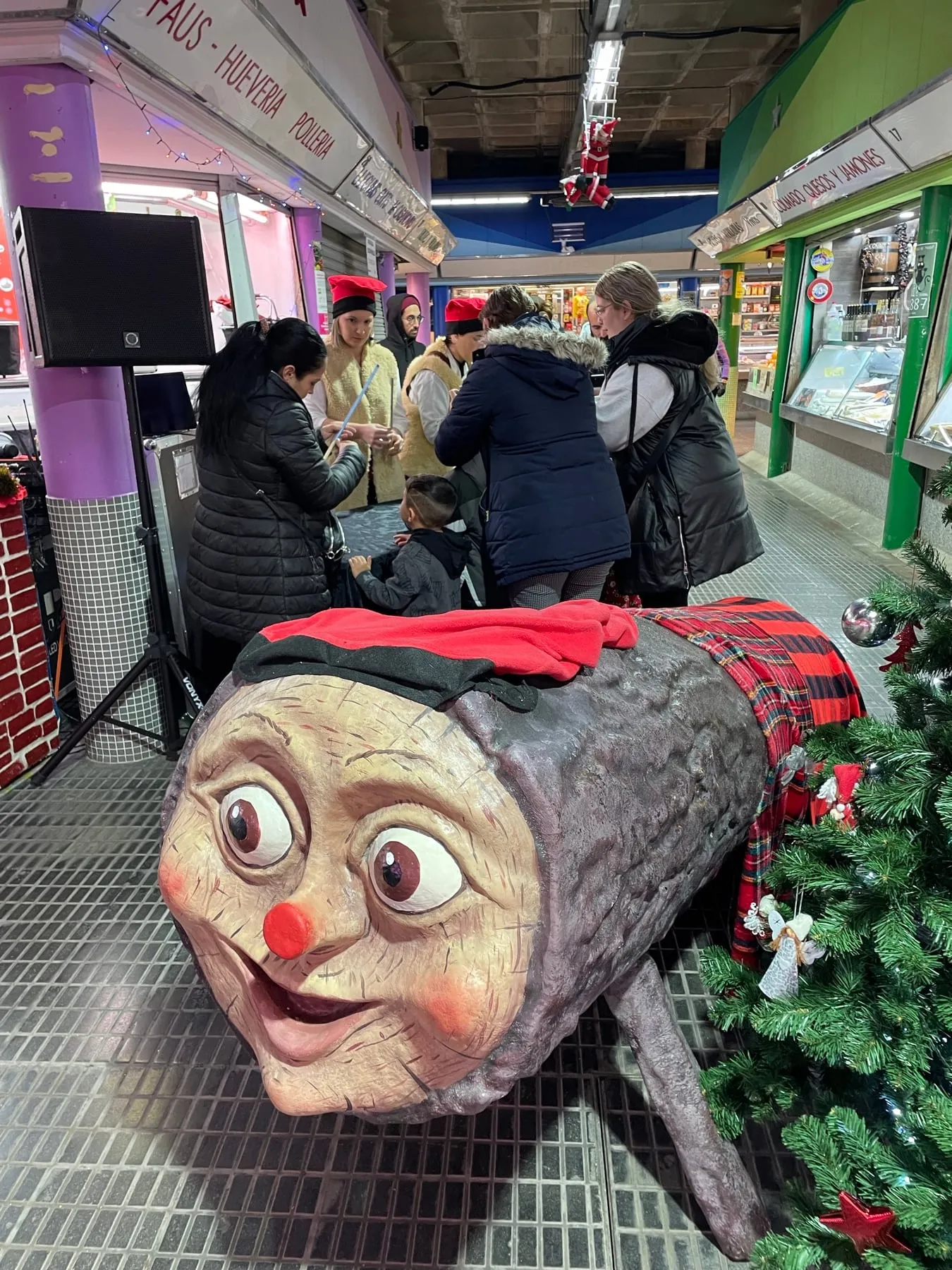 Tió gegant amb barretina i manta de quadres instal·lat en un mercat, al costat d’un avet de Nadal
