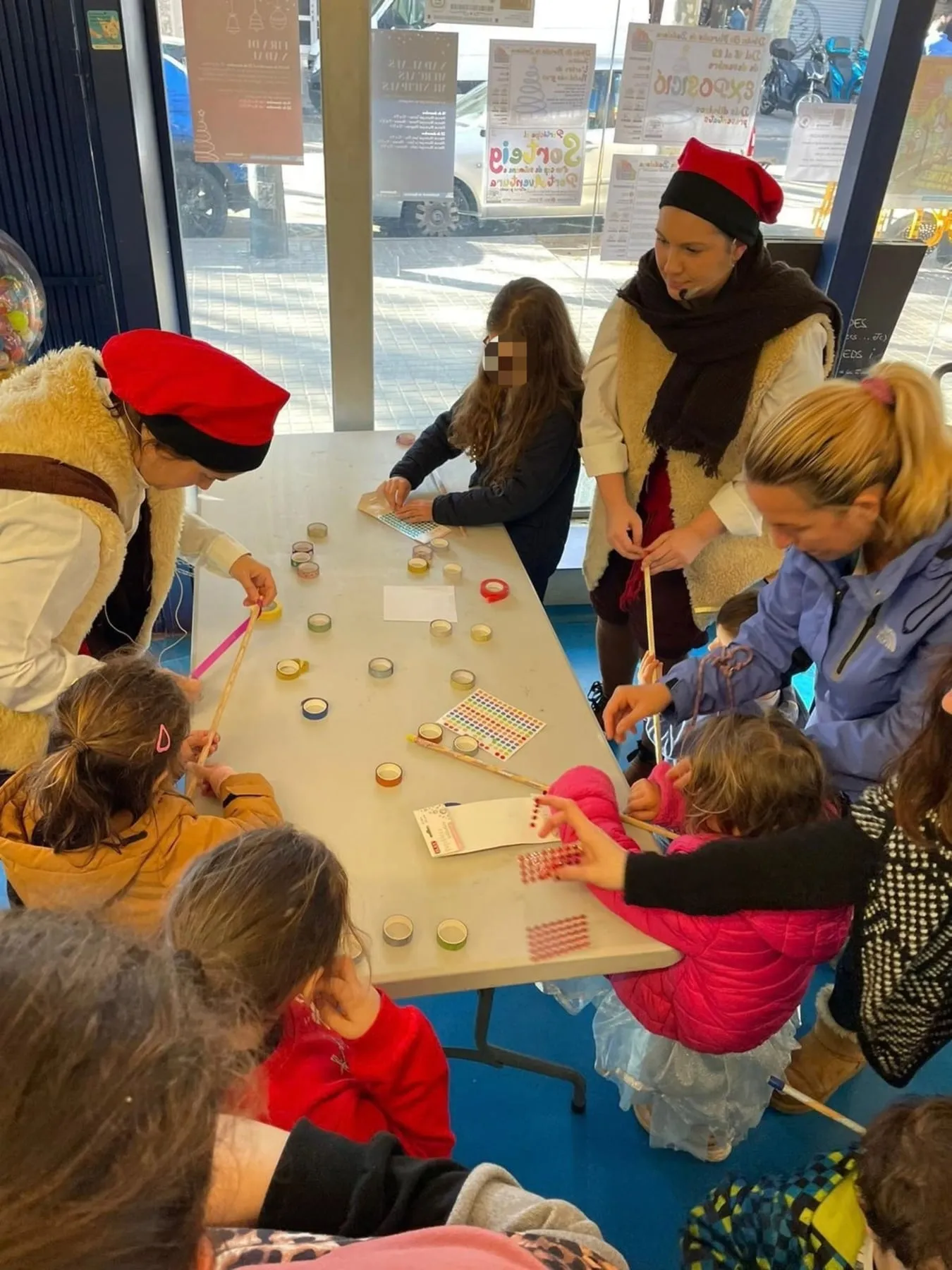 Infants decorant bastons en una taula amb cintes adhesives i gomets, ajudats per dues intèrprets amb barretina